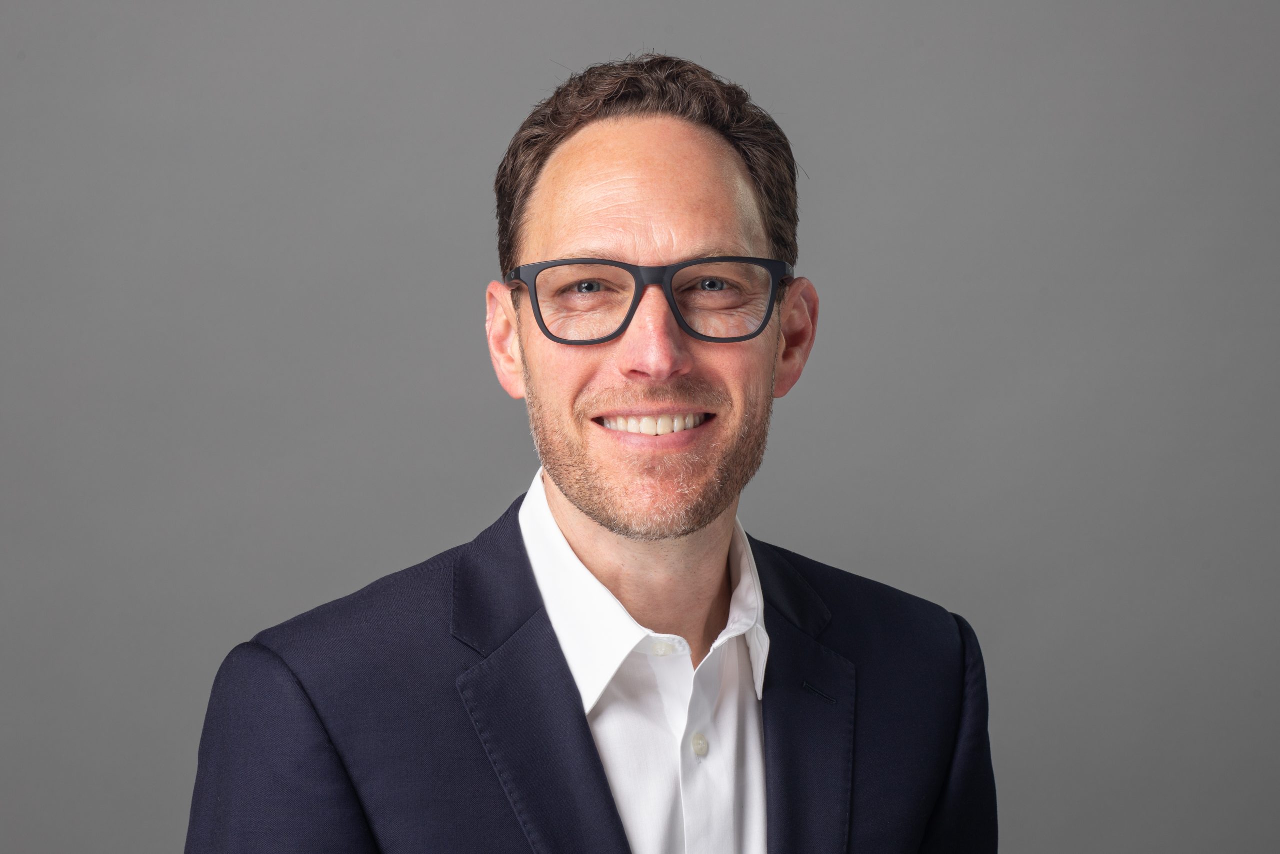 Portrait of Rob Yopps, President, smiling in a navy blazer and white shirt against a gray background.